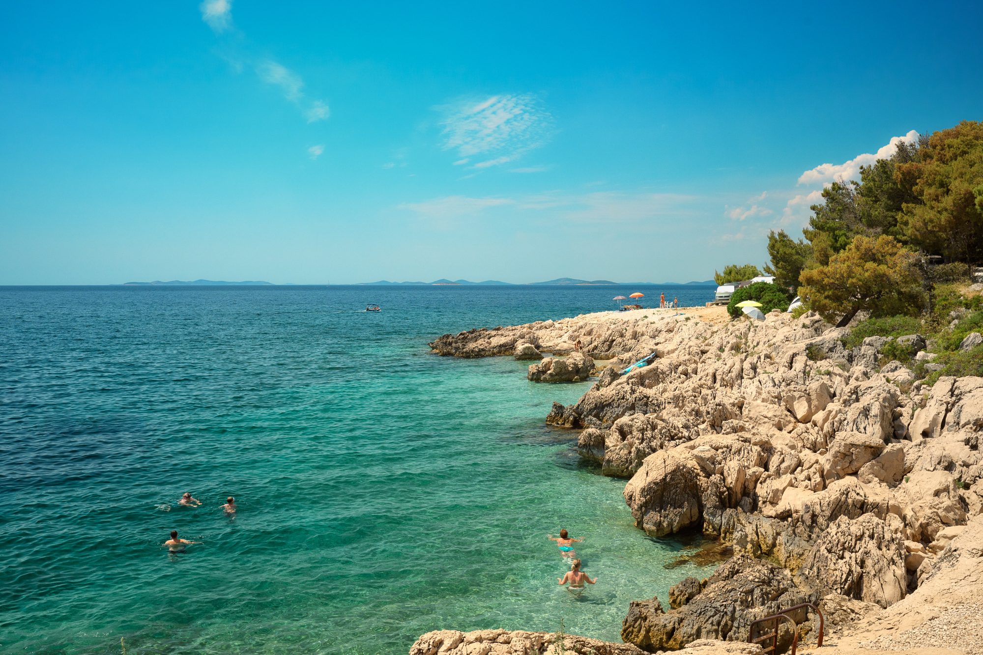 Swimmers in the clear turquoise water of a rocky Adriatic cove under a bright blue sky